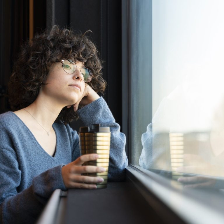 A young woman with curly hair looks out the window while holding a glass with her hand.