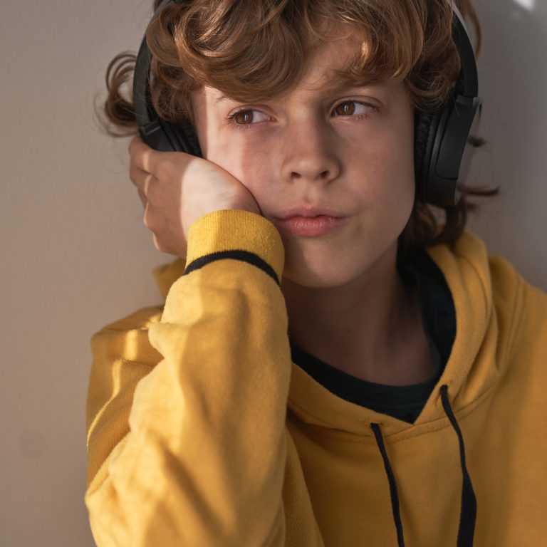 Thoughtful child in headphones and yellow sweater listening to song while relaxing alone at home and looking away